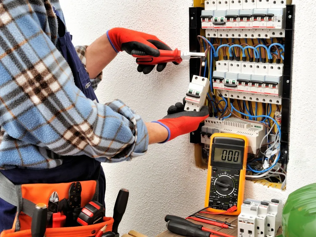 Electrician wearing red gloves uses a multimeter to test a circuit breaker panel